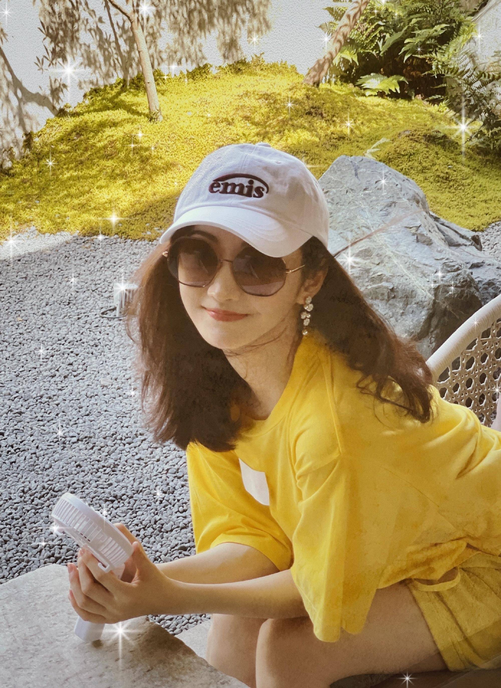 她身穿黄色衣服和短裤子,头上戴着白色的太阳帽,戴着黑超眼镜,手拿